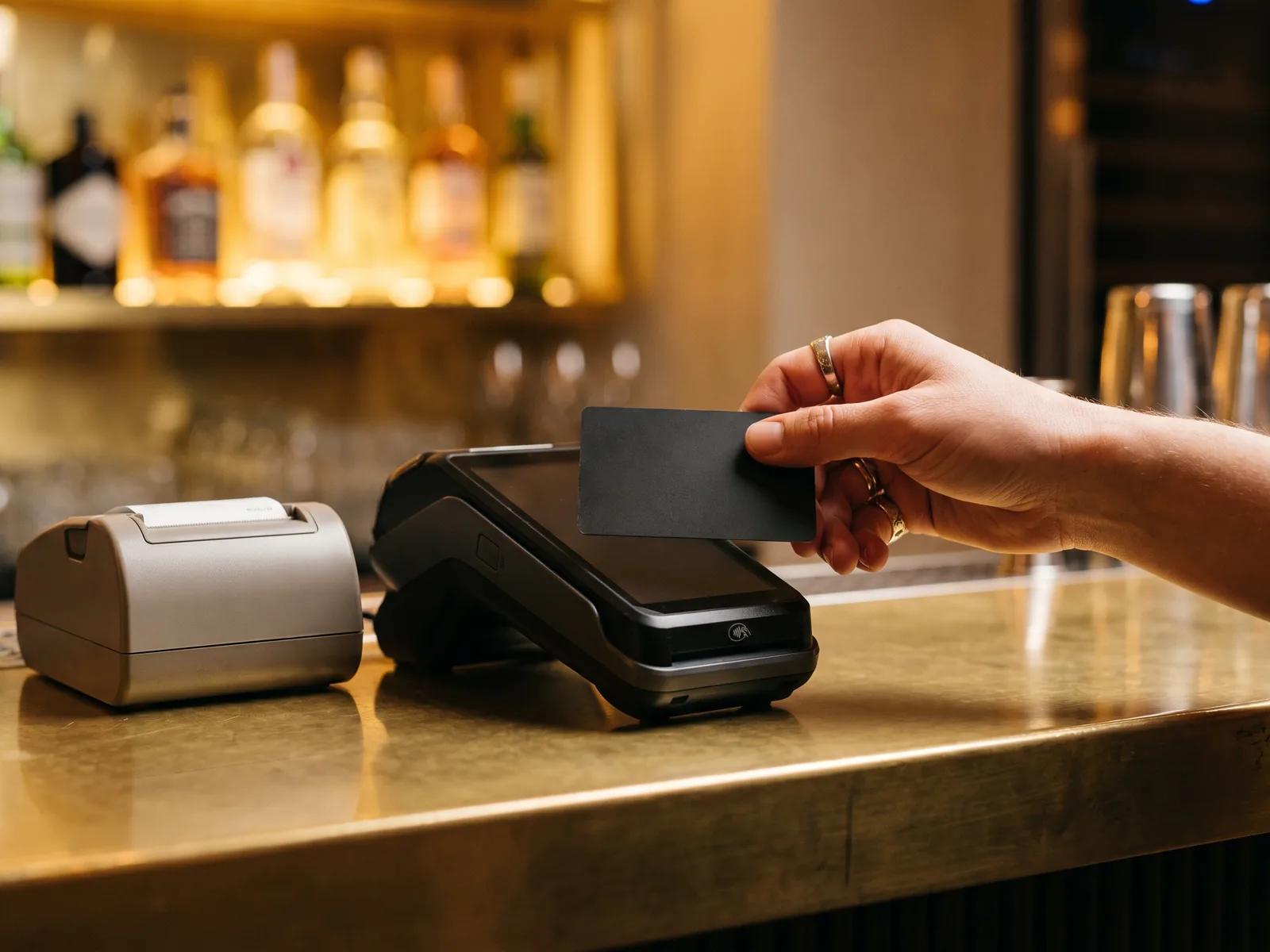 A hand tapping a card on a payment terminal at a brass bar